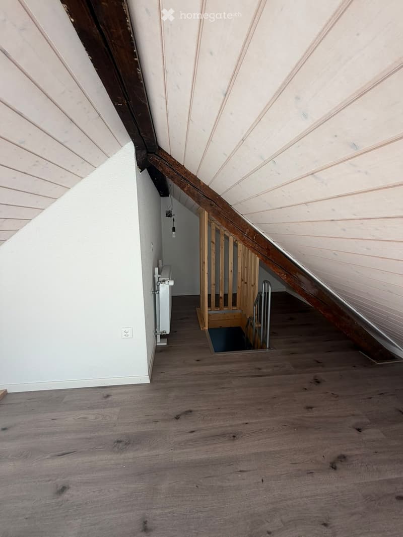 Empty attic space with wooden flooring, slanted wooden ceiling, and visible wooden beams. A small staircase leads down to another area.