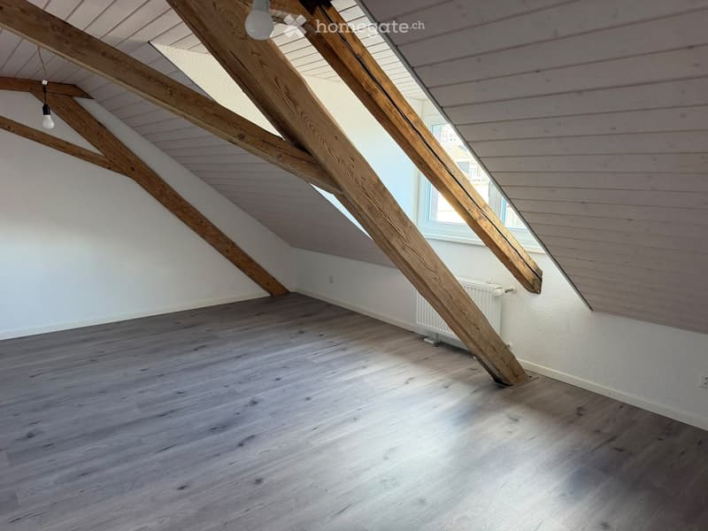 Empty attic space with wooden beams, light wooden flooring, and white walls. Natural light streams in from a window on the right side.