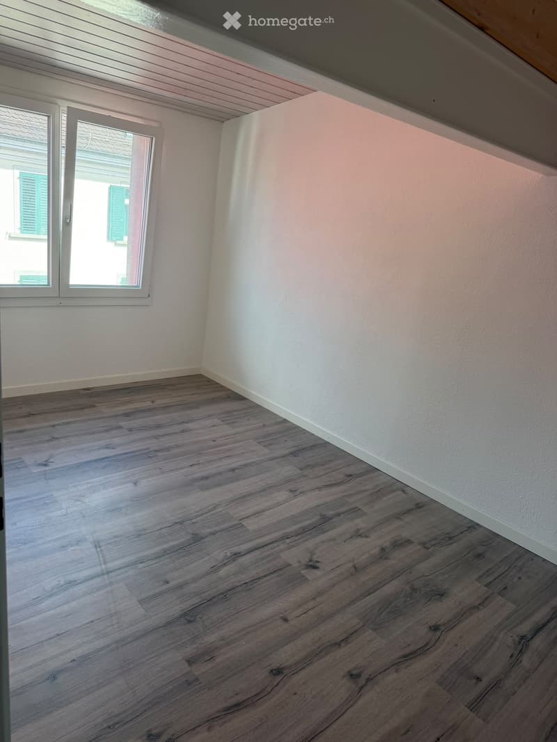 Empty room with wooden flooring and white walls, featuring a large window with shutters.