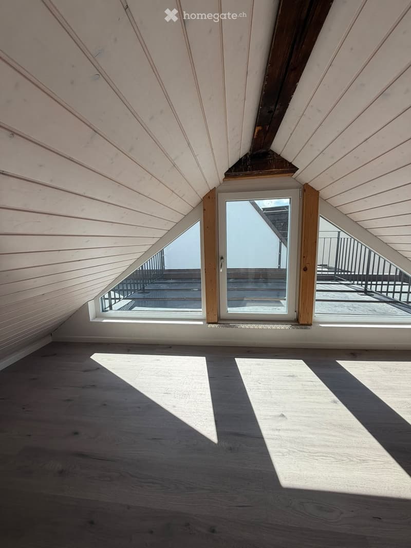 Empty attic room with wooden slanted ceilings, wooden floor, and large windows offering a view of a balcony area.