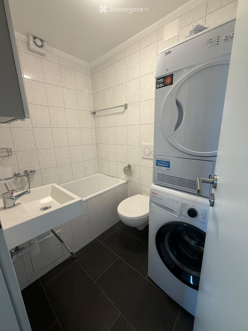 Modern bathroom with white tiled walls, black tiled floor, sink, bathtub, toilet, and stacked washing machine and dryer.