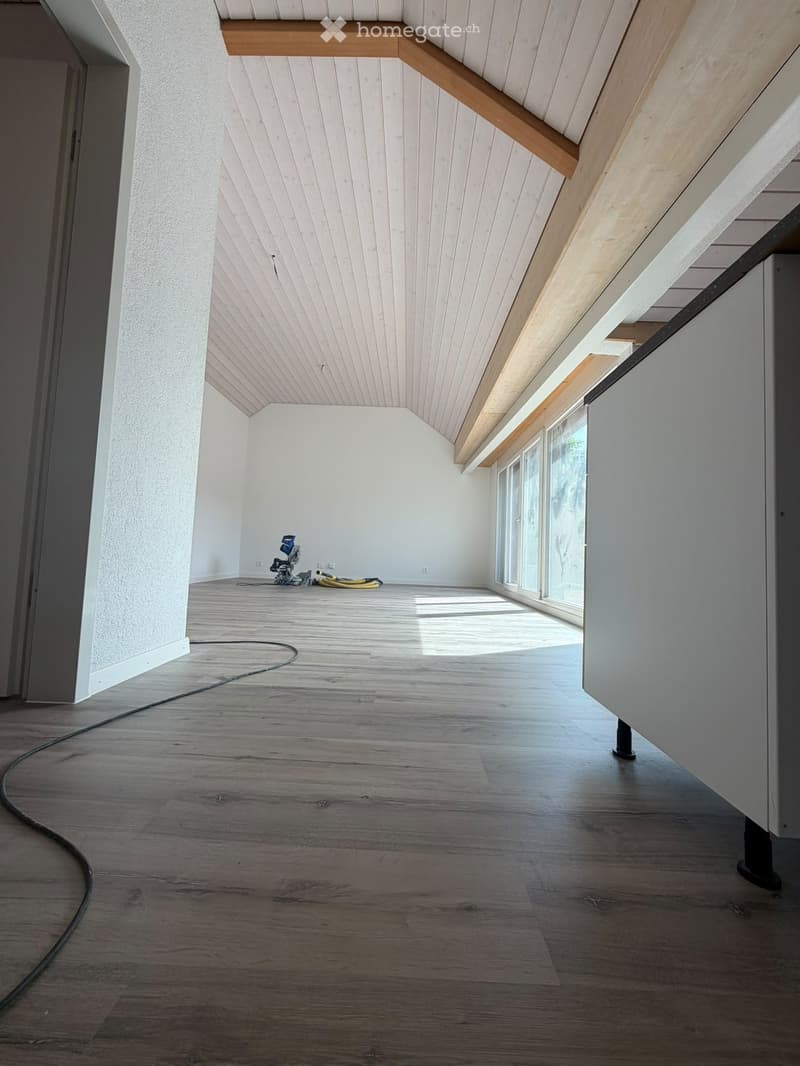 Empty room with wooden flooring, white walls, and a slanted wooden ceiling. Natural light streams in from large windows on one side.