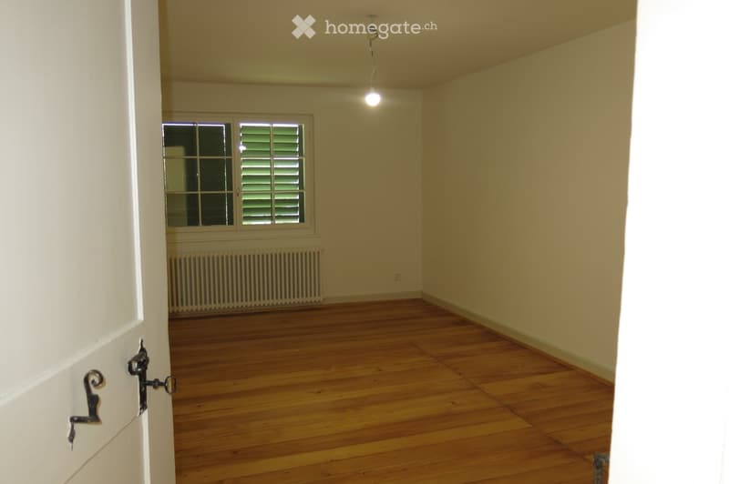 Empty room with wooden flooring, light yellow walls, a window with shutters, and a radiator below it. The room is unfurnished and has a simple, clean design.