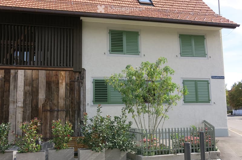 Two-story house with a tiled roof, white exterior walls, green shutters, and a wooden garage door. Small garden with plants and a fence in front.