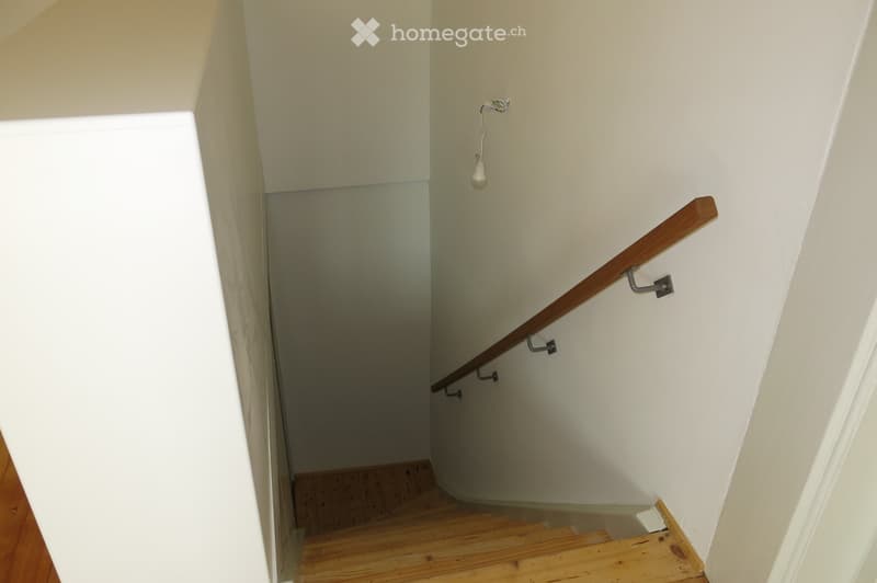A narrow staircase with wooden steps and a wooden handrail, featuring white walls and a small light fixture on the wall.