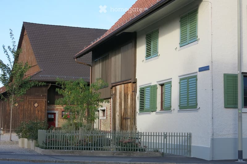 Two-story house with a red-tiled roof, white exterior walls, green shutters, and a wooden garage. A small garden with a fence is visible in front.