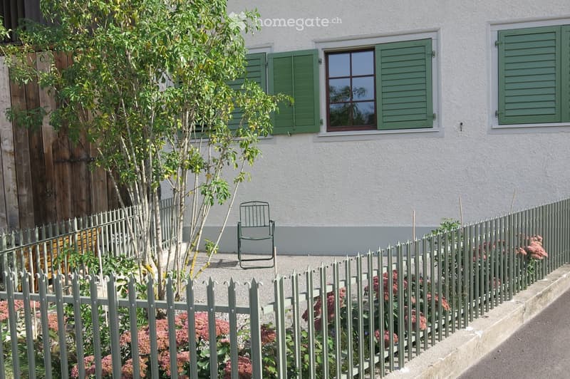 A small garden area with a white fence, green shutters, a small tree, and a folding chair placed near the house.