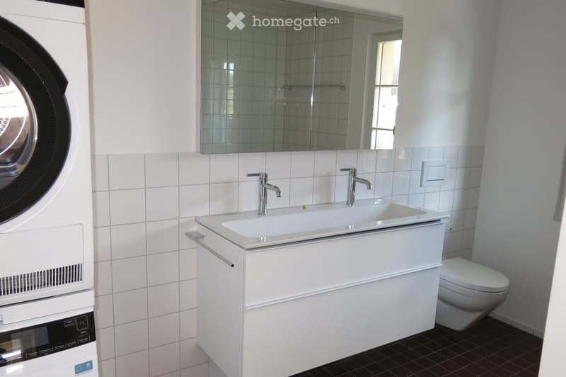 Modern bathroom with white tiles, a double sink with chrome faucets, a large mirror, a toilet, and a washing machine.