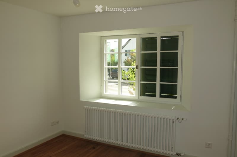 Empty room with wooden flooring, white walls, a radiator, and a large window offering a view of the street and greenery outside.