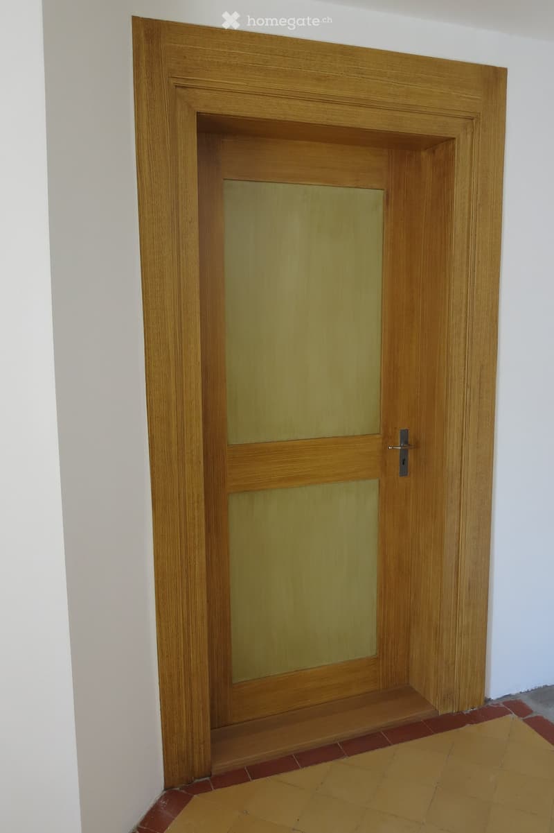 Wooden door with frosted glass panels, set in a white-walled corridor with tiled flooring in red and beige.