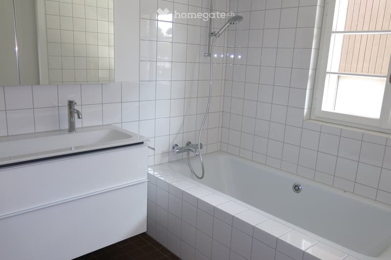 Modern bathroom with white tiled walls and floor, featuring a bathtub with a handheld shower, a sink with a modern faucet, and a large mirror above the sink. A window provides natural light.