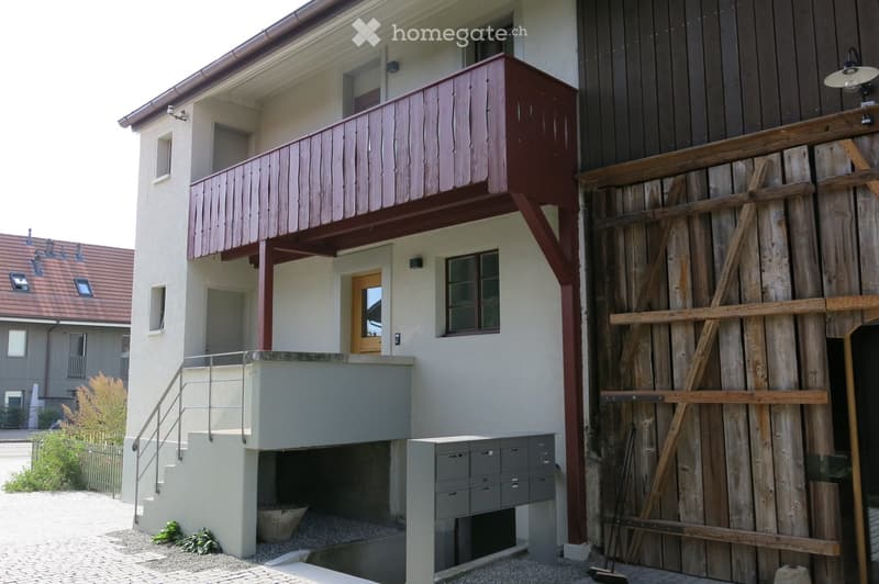 Modern two-story building with a raised ground floor, balcony, and wooden shutters on the upper level.