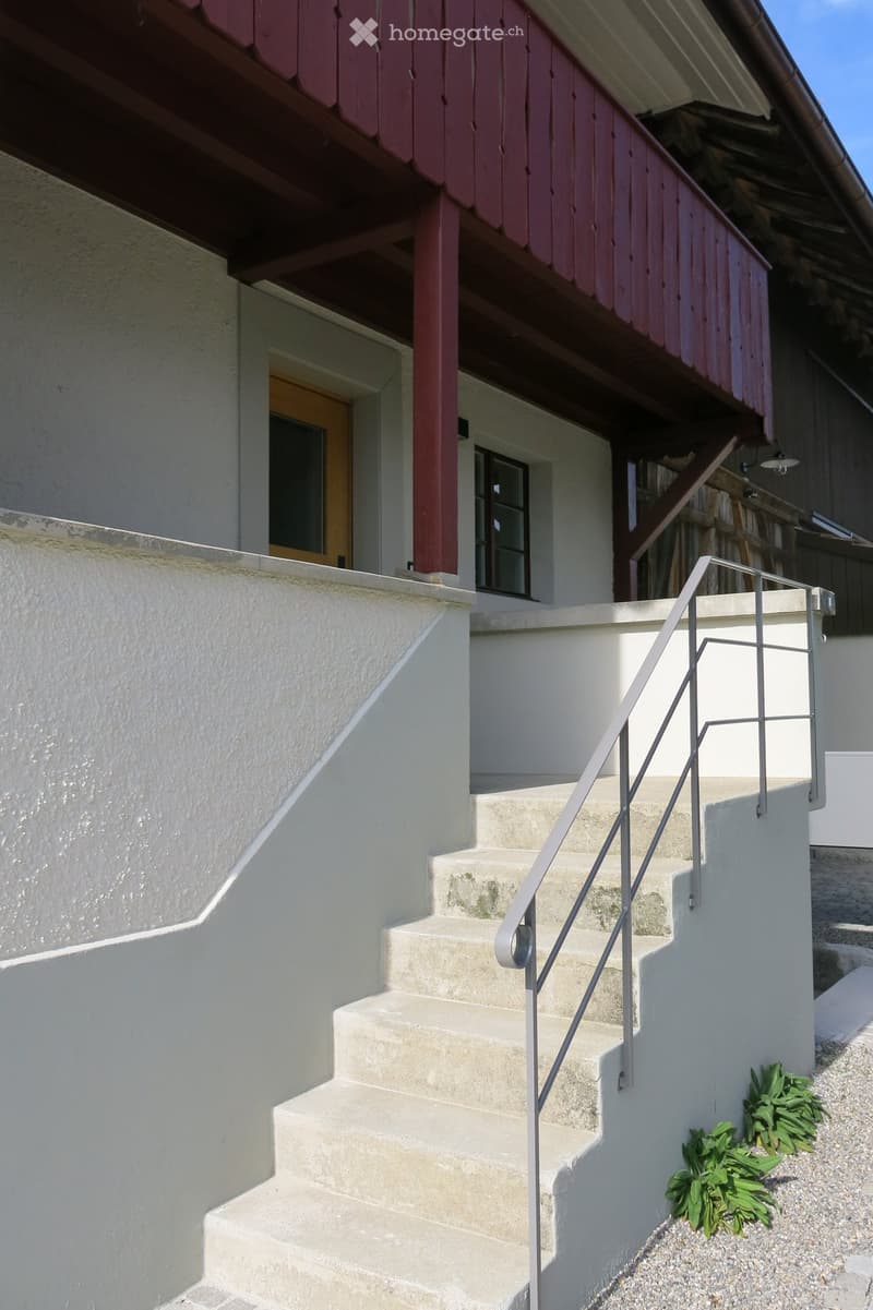 Exterior view of a two-story house with a raised ground floor, featuring a staircase with a metal railing leading up to the entrance. The house has white walls, a wooden balcony with red trim, and a small garden area with green plants near the base of the