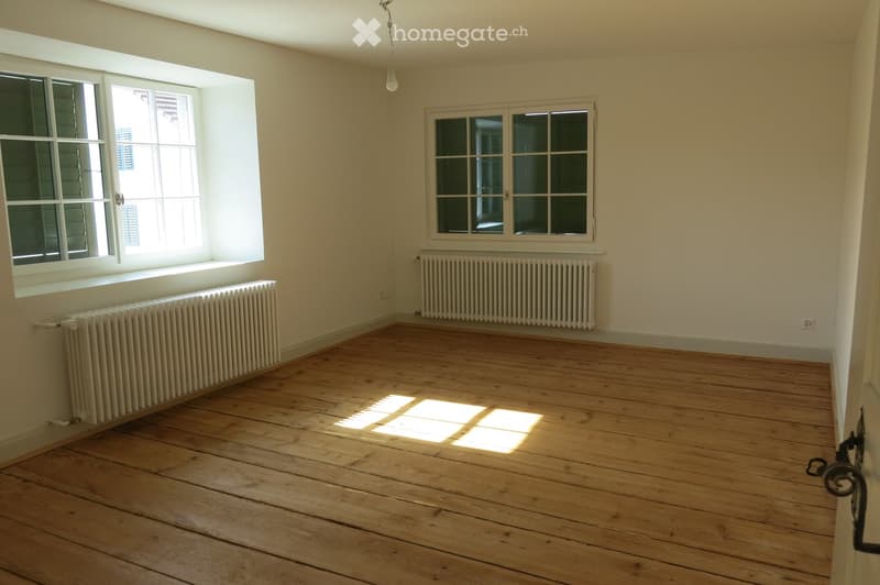 Empty room with wooden flooring, two windows, radiators, and natural light streaming in.