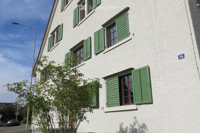 Two-story house with white exterior walls, green shutters, and multiple windows. The house number "54" is displayed on the wall.