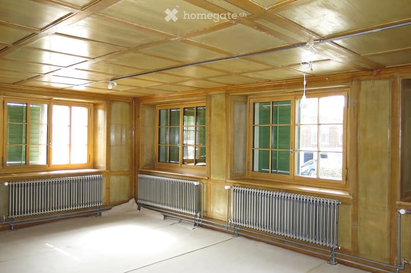 Empty room with wooden panel walls and ceiling, large windows with green shutters, radiators along the walls, and a hanging light fixture.