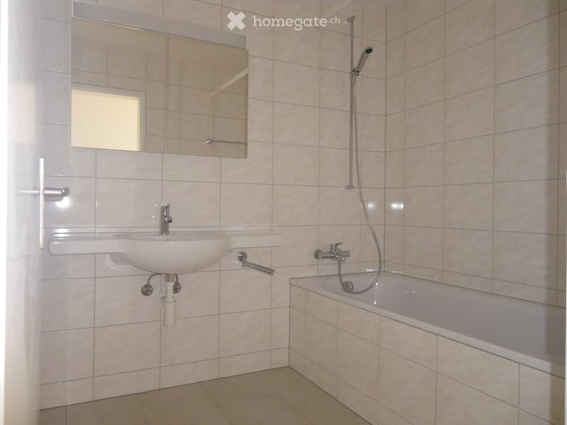 Modern bathroom with a white sink, bathtub, and a large mirror above the sink. The walls and floor are tiled in a light beige color.