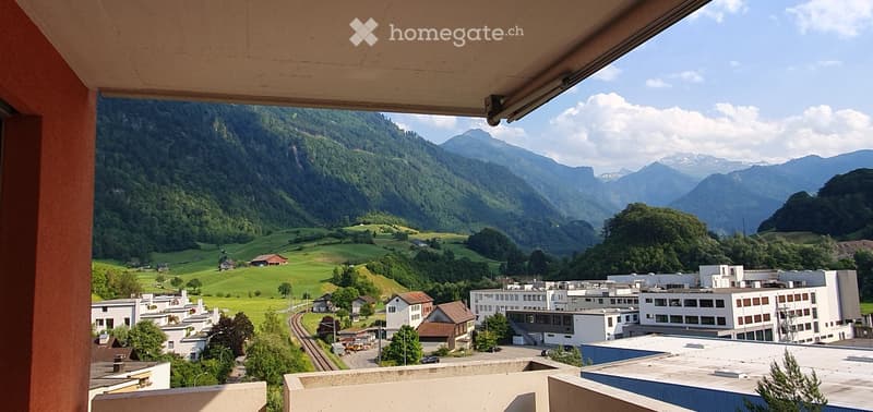 Balcony overlooking a scenic valley with green hills, scattered houses, and distant mountains under a partly cloudy sky.