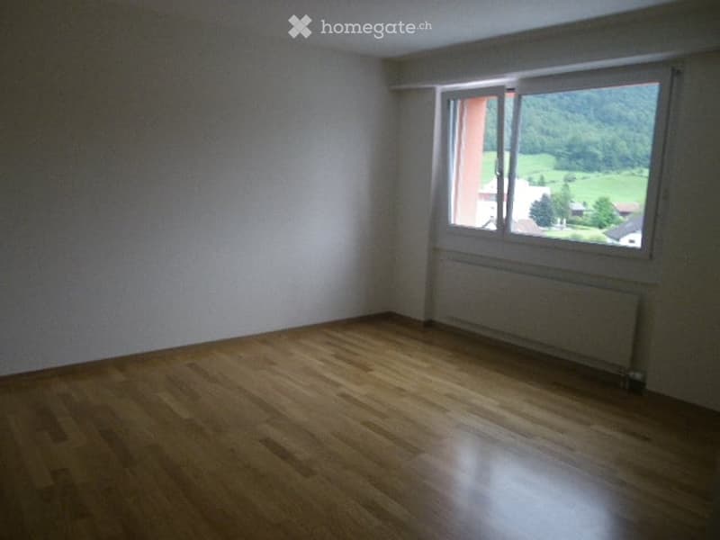 Empty room with wooden flooring, white walls, and a large window offering a scenic view of greenery and a house outside.