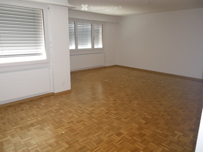 Empty room with wooden parquet flooring, white walls, and large windows with blinds. No furniture or equipment present.
