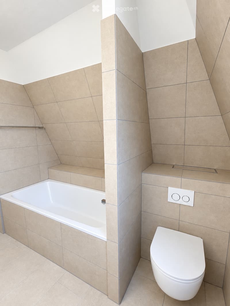 Modern bathroom with beige tiled walls and floor, featuring a bathtub and toilet, with a sleek design and built-in shelves.