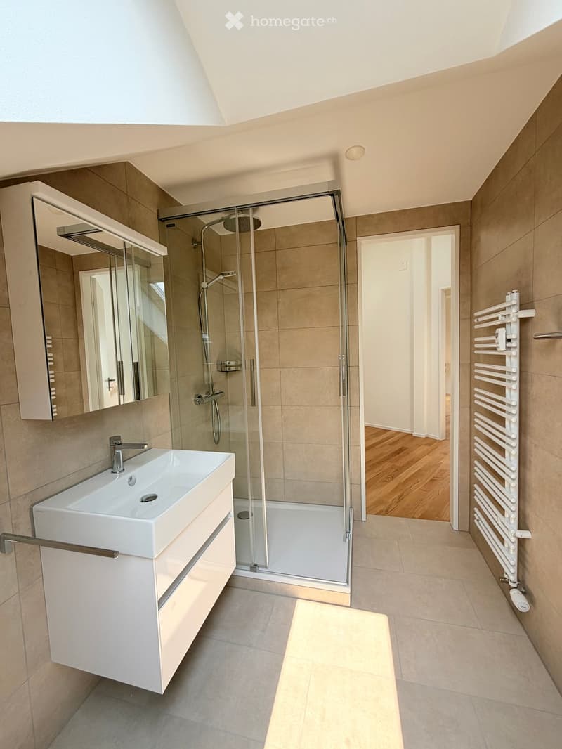 Modern bathroom with a white sink, large mirror, glass-enclosed shower, and towel rack. Light-colored tiles and wooden flooring are visible.