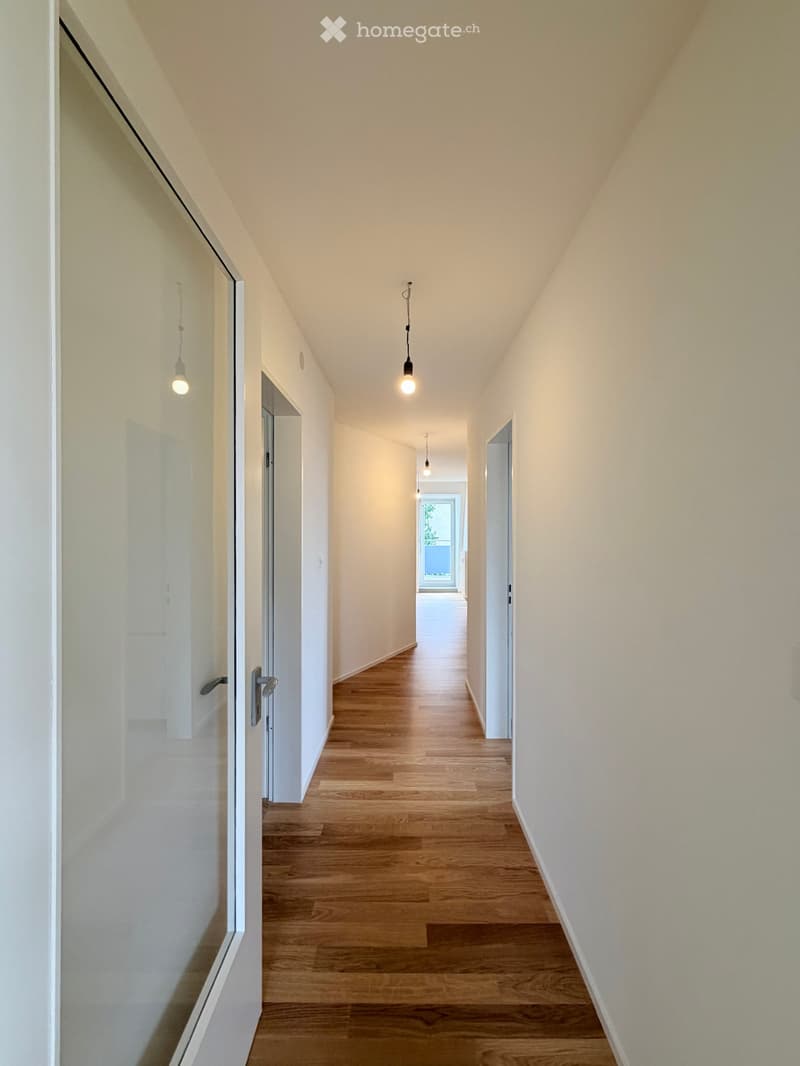 A clean, modern hallway with light wooden flooring, white walls, and several pendant lights hanging from the ceiling. Doors are visible on both sides, leading to other rooms.