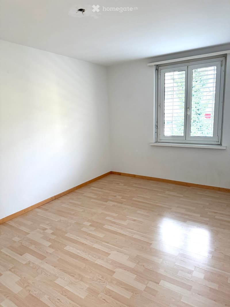 Empty room with light wooden flooring and white walls, featuring a large window with blinds on the right side.