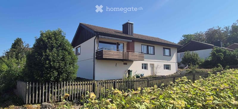 Two-story house with a balcony, surrounded by greenery and a wooden fence.