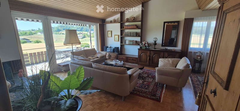 Bright living room with large windows offering a scenic view, featuring a beige sofa, coffee table, armchair, table lamp, bookshelf, mirror, flower arrangement, and a small table with chairs near the window.