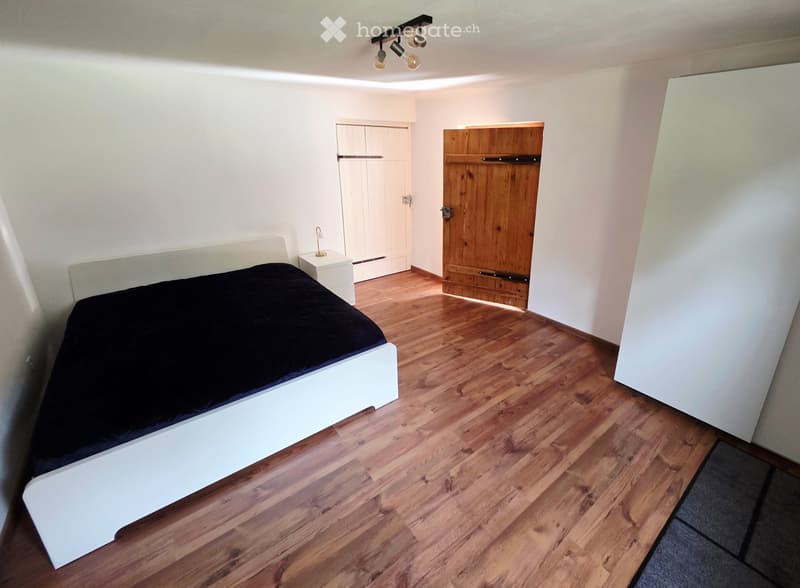 Modern bedroom with a white bed, dark bedding, wooden floor, white nightstand, and a lamp. Two doors are visible, one white and one wooden.