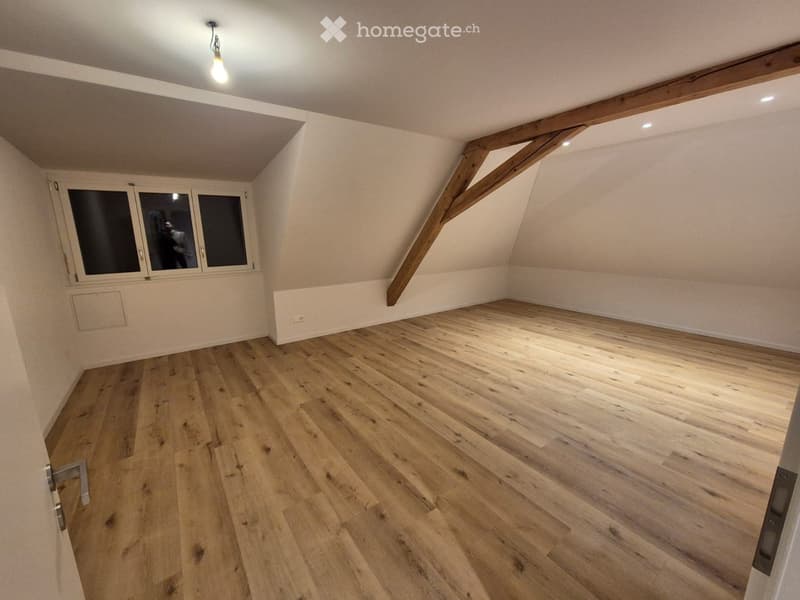 Empty attic room with wooden flooring, exposed wooden beams, and large windows.