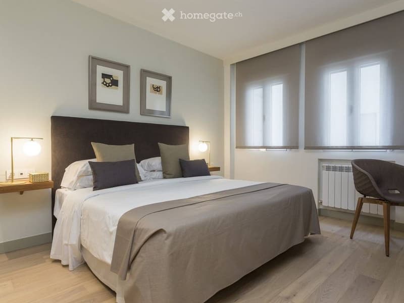Modern bedroom with a large bed, nightstands, a chair, and two picture frames on the wall. The room features wooden flooring and large windows with blinds.