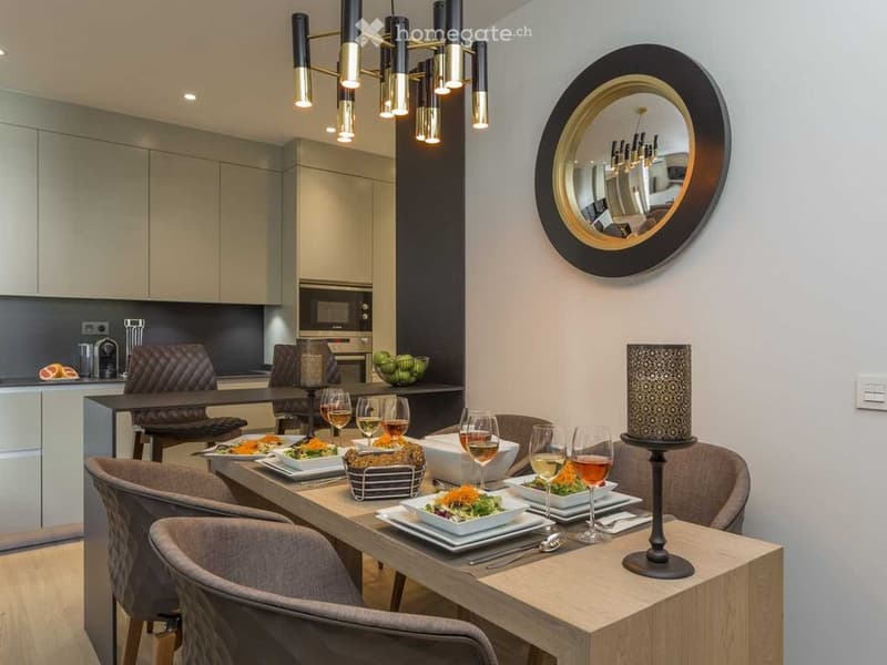 Elegant dining area with a wooden table set for a meal, surrounded by modern chairs, wine glasses, and decorative plates. A sleek lamp and a round mirror adorn the wall, with a contemporary kitchen area visible in the background.