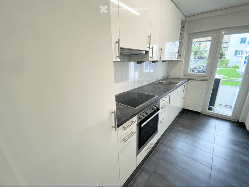 Modern kitchen with white cabinets, dark countertops, induction cooktop, oven, and sink. Large window offers a view of the garden.