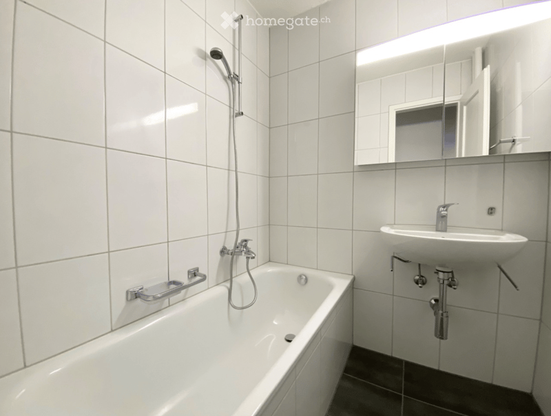 Modern bathroom with white tiled walls and floor, featuring a bathtub with a handheld shower, a sink with a modern faucet, and a large mirror with built-in lighting.