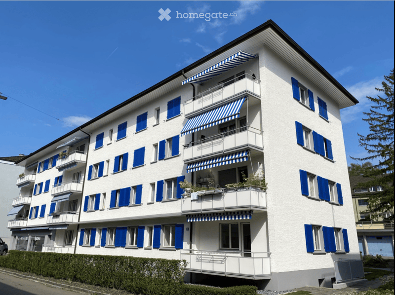 A multi-story white building with blue shutters and balconies, featuring striped awnings and potted plants on the balconies.