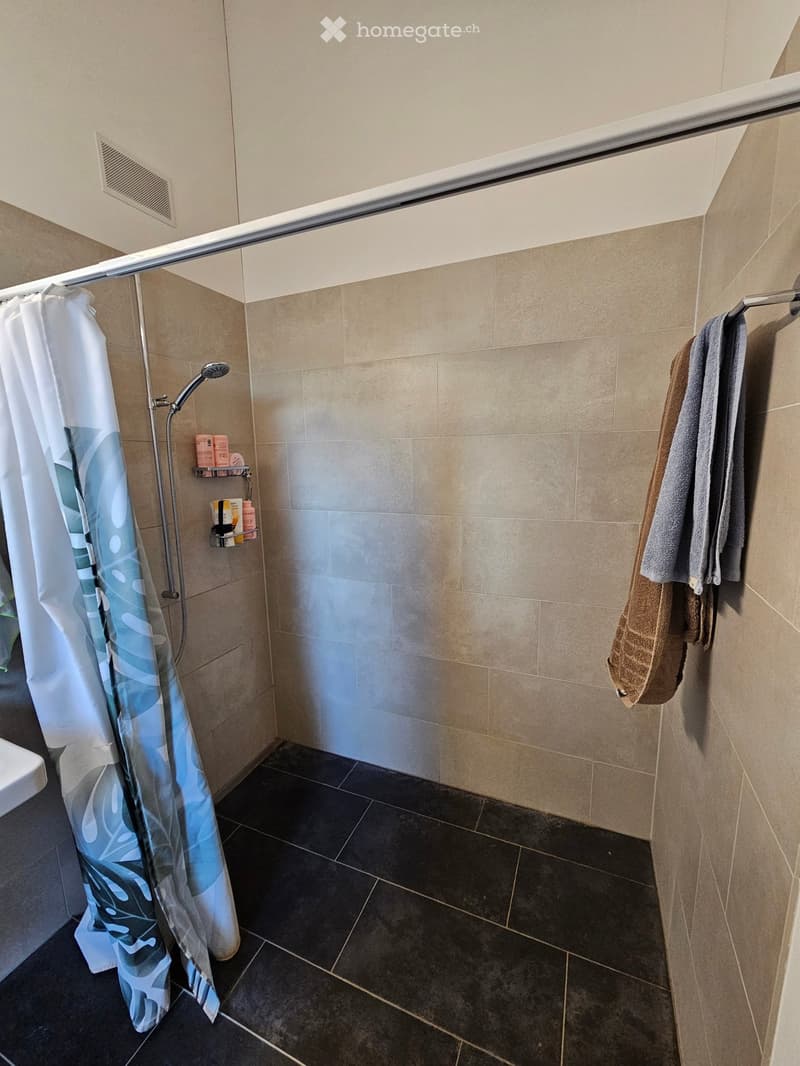 Modern bathroom with a walk-in shower, beige tiles, dark floor tiles, a white shower curtain, a towel rack with towels, and a shower shelf with toiletries.