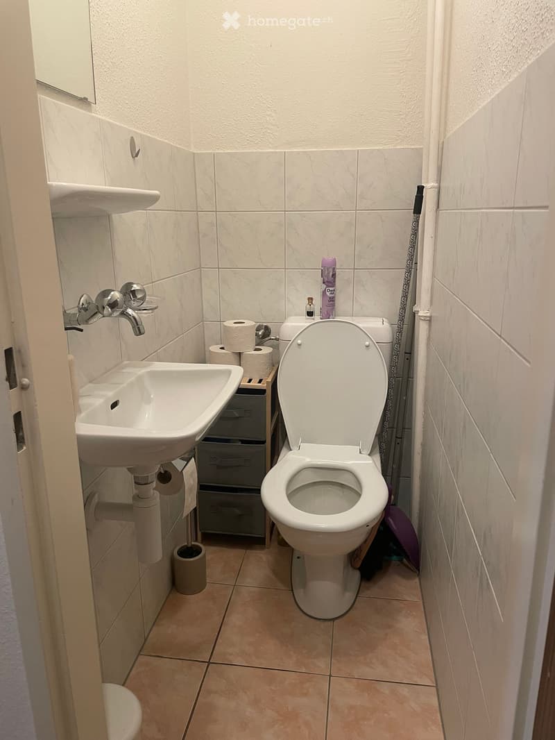Small bathroom with a white sink, chrome faucet, toilet with lid up, toilet paper holder, and a bottle on the toilet tank. Beige tiled floor and walls.