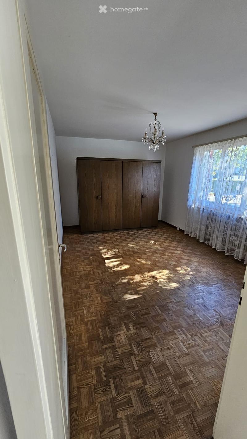 Empty room with wooden herringbone flooring, white walls, a large wooden wardrobe, and a chandelier hanging from the ceiling. Natural light filters in through sheer curtains on the window.