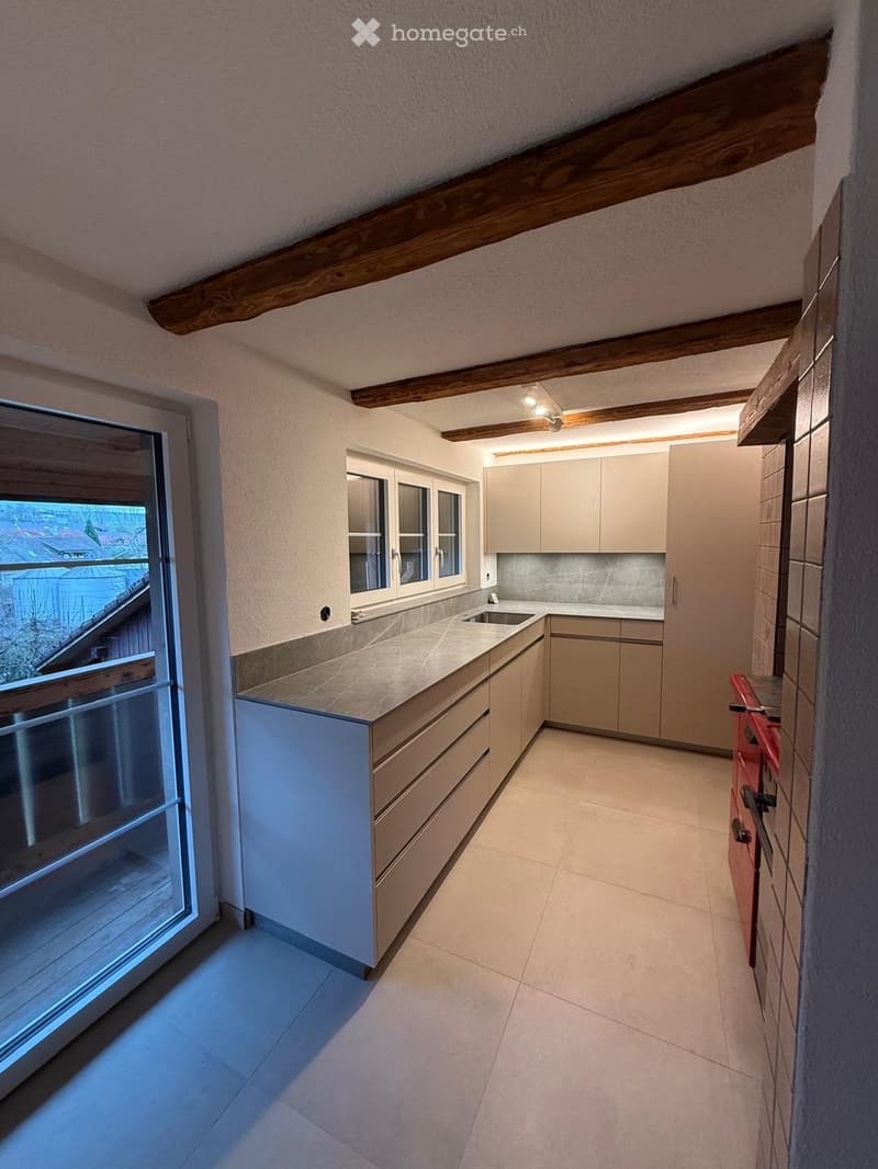 Modern kitchen with light-colored cabinets, a central island, and a sink. The space features exposed wooden beams on the ceiling and large windows providing natural light.