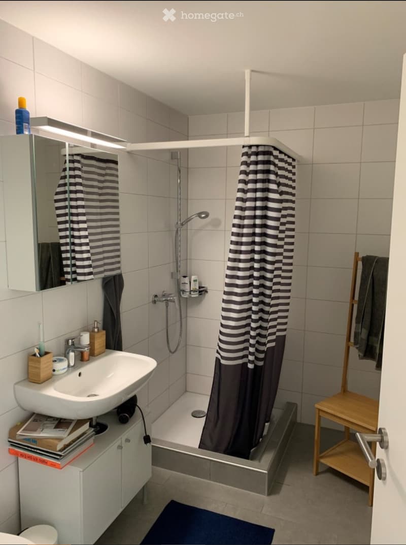 Modern bathroom with a white sink, mirror, bathtub with a black and white striped shower curtain, a small wooden stool, and various toiletries on the sink counter.