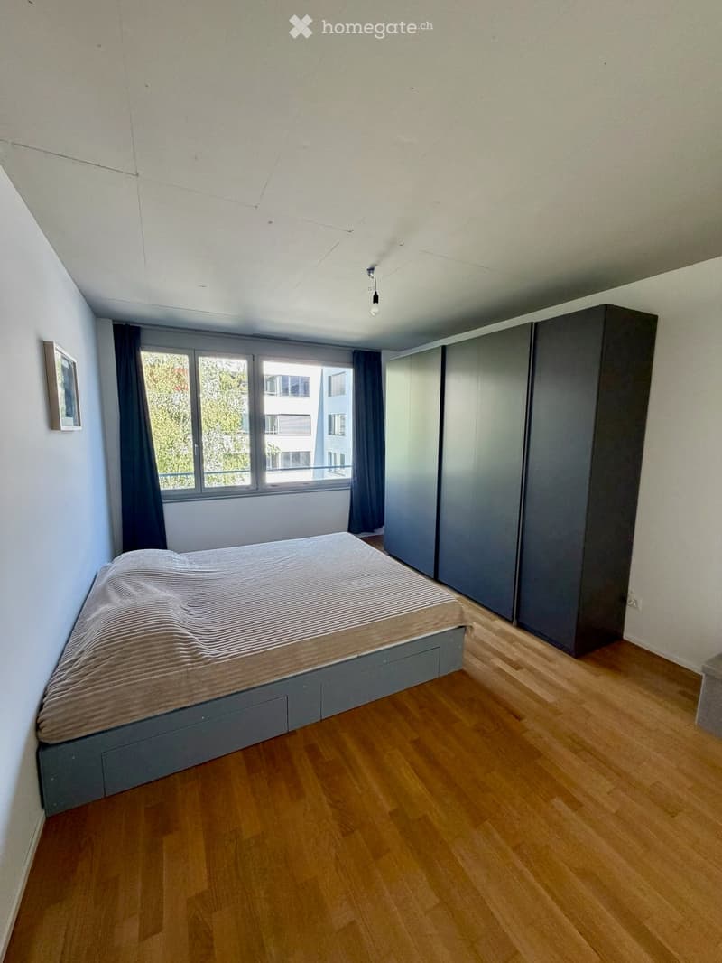 Modern bedroom with wooden flooring, large window with curtains, grey bed with striped bedding, built-in wardrobe, and a picture frame on the wall.