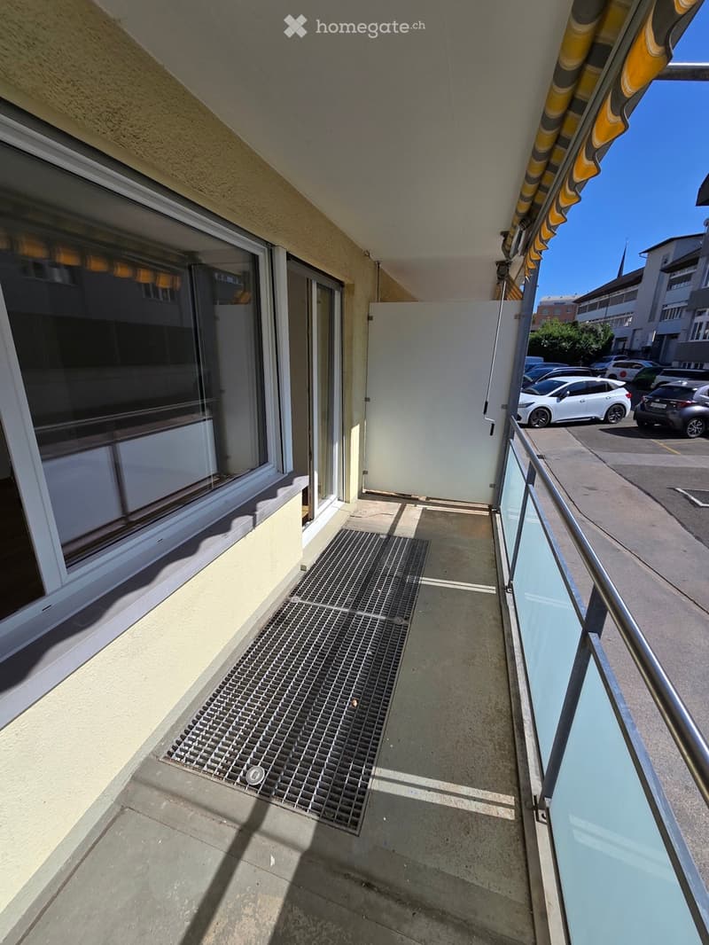 Balcony with metal grid floor, glass railing, and a view of a parking lot with several cars.
