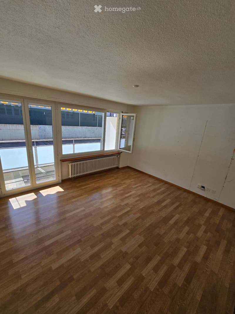 Empty room with wooden flooring, large windows, radiator, and a small wooden bench near the windows.