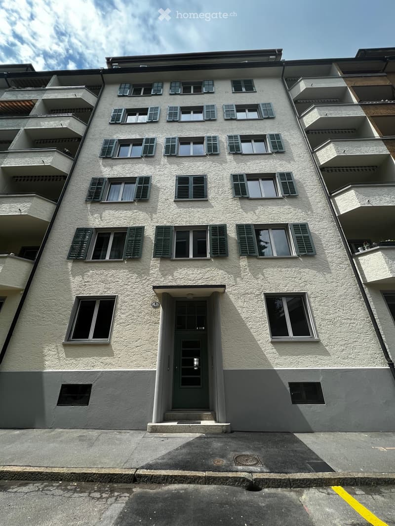 Four-story residential building with light beige textured facade, multiple windows with green shutters, and a central entrance door.