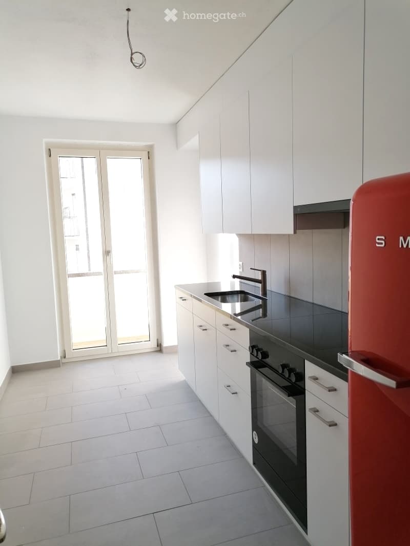 Modern kitchen with white cabinets, black countertops, a sink, faucet, oven, and a red refrigerator. Features a large window with a view.