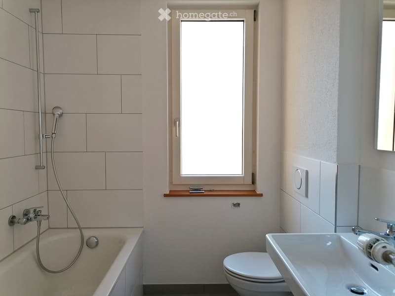 Modern bathroom with a bathtub, toilet, sink, and showerhead. Light-colored tiles and a window provide natural light.