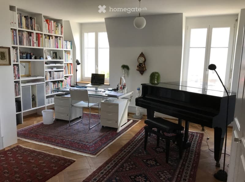 Brightly lit room with wooden flooring, large windows, bookshelf filled with books, desk with laptop, chair, piano, and decorative items.