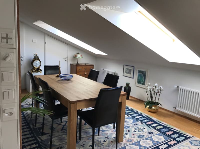 Bright attic dining area with wooden dining table, black chairs, decorative lamp, vase with flowers, and artwork on walls.